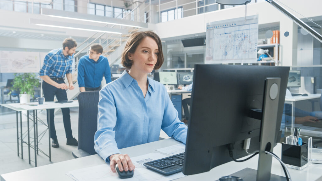 Frau in blauem Hemd sitzt vor einem Computerbildschirm und arbeitet, im Hintergrund stehen 2 Männer an einem Tisch und reden miteinander